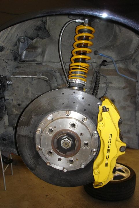 997 Turbo upgrade to 9e 28 by Nine Excellence (pic heavy) - Page 2 - Porsche General - PistonHeads - The image shows a close-up of the underside of a car, focusing on a disc brake with track pads. There is an extended yellow coil spring partially visible under the car, which is likely associated with the suspension. The brake disc is attached to a hub assembly, which includes circulating bolts to secure the brake pad. Adjacent to the pads, a brake caliper is visible, with the brand name "RB6" inscribed on it. The environment appears to be a garage or a workshop area as indicated by the visible suspension components and the presence of a black and yellow tire.