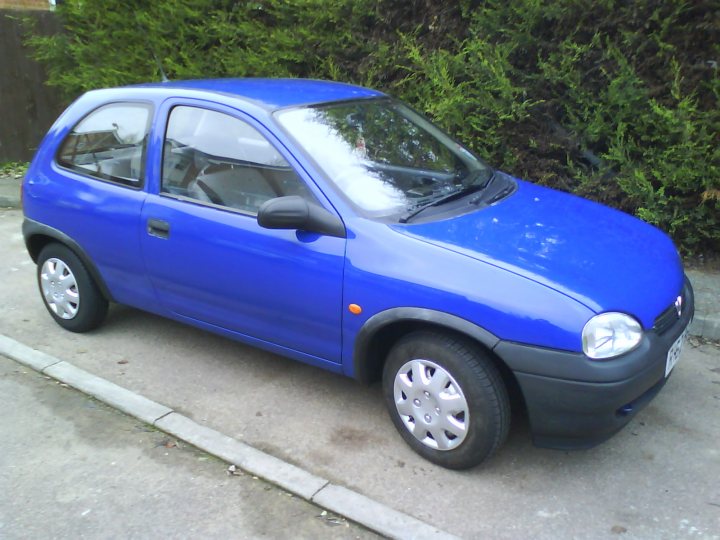 Pistonheads Wheels Hat Ready Corsa Tin - The image captures a scene of a small blue car parked on the side of a road or driveway. The car, with its compact size and distinctive blue color, is the most prominent object in the image. The setting appears to be outdoors, with the car positioned close to a fence and vegetation, providing a natural backdrop to the vehicle. The surface beneath the car is paved, indicating an urban or suburban location, which is further supported by the presence of the fence and vegetation.