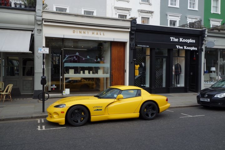 Black OEM wheels - Page 1 - Vipers - PistonHeads - The image showcases a vibrant city street lined with shops. The most striking feature in the picture is a **yellow convertible sports car** parked on the street right in front of a shop named "**The Kooples**". This sports car has a **black hood** and a **black soft top** that adds to its unique appearance. 

In the foreground, there's a parking meter and a black car parked behind the yellow sports car. The street appears to be relatively quiet at the moment, with no people visible in the image. 

The background reveals the full length of the city street with various shops and buildings. The architectural style of the buildings suggests an urban setting, possibly in a part of town that is well-known for its shopping.