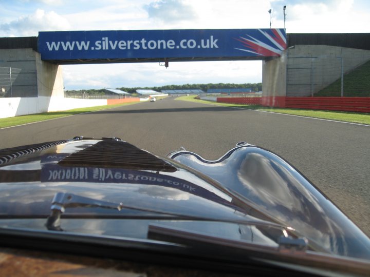 Head Fixed Coupe Pistonheads - The image showcases a Formula 1 race car from the rear, captured during the Dunlop tire test at the Silverstone Race Circuit. The car, stripped of its windscreen and covered in Dunlop's signature branding, is shown entering the track under an overhead archway displaying the circuit's name. The track itself is clearly marked, with a fencing line visible along the edge and multiple lanes of the track. The car's position and the overcast sky suggest it is either preparing for its run or just concluding it. The image conveys a sense of motion and the intensity of the sport.