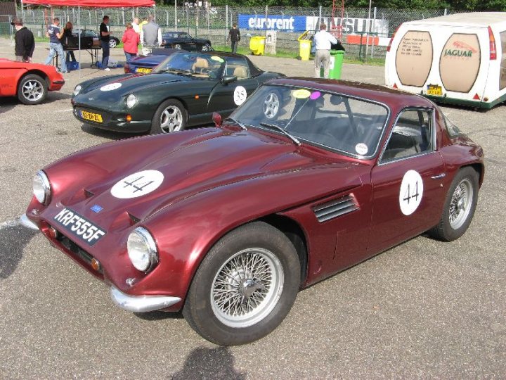 Early TVR Pictures - Page 126 - Classics - PistonHeads - The image showcases an outdoor parking lot scene where a classic red sports car is the highlight. The car prominently features the number 4 and a white decal with the letter "H" on its front and side. It shares the lot with other classic cars exhibited in a line. Behind the cars, there is a white van with the word "Jaguar" on it. In the background, there are people walking around, and a few tents are set up, suggesting an event or car show might be taking place. A garbage bin is also visible in the far left of the image.