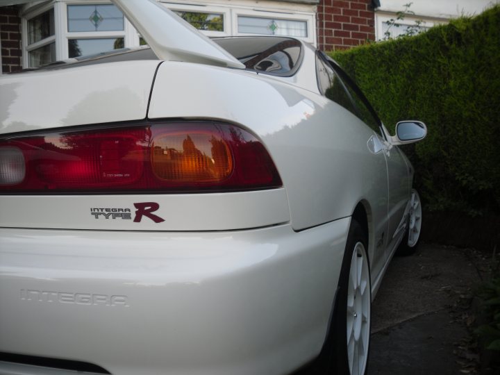 Pistonheads - In the image, there is a light blue or white car parked on a driveway next to a house. The car is facing the viewer with its trunk open, revealing the back side of the vehicle. It's a sunny day, as suggested by the natural light illuminating the scene. The car features a distinctive red emblem on the case, adding a pop of color to the otherwise neutral hues. The backdrop of the house and driveway provides a typical residential setting.