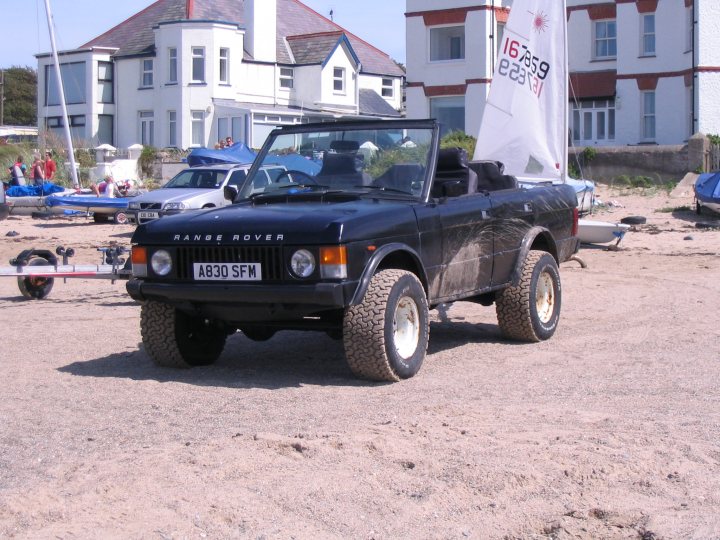 Estate Version Pistonheads - The image shows a black Range Rover Sport with a white sail attached to the roof, parked on a sandy area covered with debris. The car has tinted windows and is equipped with chunky off-road tires. In the background, there's an abandoned white sailboat on the sand. A house with white walls and a red roof can be seen in the distance, along with some other vehicles and elements indicative of a beach or seaside location.