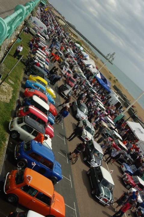 Pistonheads Brighton London - The image displays a large parking lot filled with a variety of cars in various colors and models. People are scattered among the vehicles, some walking and others standing. In the background, numerous umbrellas are opened on the ground, likely providing shade or marking spaces. The parking lot is bordered by a grassy area and extends towards a body of water. The sky overhead is cloudy, and there are no visible texts or signage in the image.