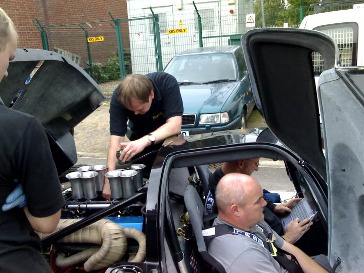 Ultima Pistonheads Track Maximise - The image captures a moment inside a large sports car with its hood open. A man, dressed in a black shirt, stands next to the car, interacting with the engine. The interior of the car features racing-grade seats and a steering wheel, suggesting a high-performance vehicle. Another man, wearing a shirt with a visible logo, is seated in the car, looking out of an open window. The car itself is sleek and impressive, with a vibrant blue color matching the man's shirt. In the background, there's a glimpse of a town setting, which provides context for the location of this scene.