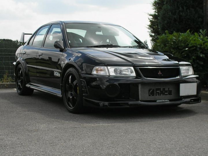 Registeredpics Evo Pistonheads Newley - The image depicts a black sedan parked in a lot. The car appears to be a Mitsubishi Galant, as indicated by the badge on the front. The vehicle is equipped with alloy wheels and is also fitted with a greenish-blue air intake. The car is positioned perpendicular to the viewer, showing the side profile with its black finish. The backdrop of the image features a green fence, suggesting an outdoor, possibly private, parking area.