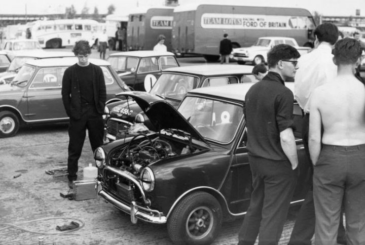 A 'period' classics pictures thread (Mk III) - Page 13 - Classic Cars and Yesterday's Heroes - PistonHeads UK - This is a black and white photograph capturing an outdoor scene of people standing around cars, which are parked on a surface. In the center of the image stands a person who appears to be examining one of the vehicles closely. The people are dressed casually, with some visible facial features such as hairstyles and glasses. There are several car models present, including a vintage convertible in the foreground and other cars scattered around in the background. In the distance, there's a truck parked, adding to the overall atmosphere of a gathering or event involving vehicles.