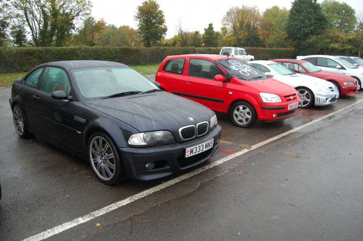 Association Castle Stroke Pistonheads Combe Day Supercar - The image depicts a parking lot filled with cars, predominantly older models. In the center of the frame stands a red Volkswagen Lupo, parked evenly between two other vehicles, one black and one silver. The Lupo's position in the lot, and its striking red color, make it the focal point of the image.