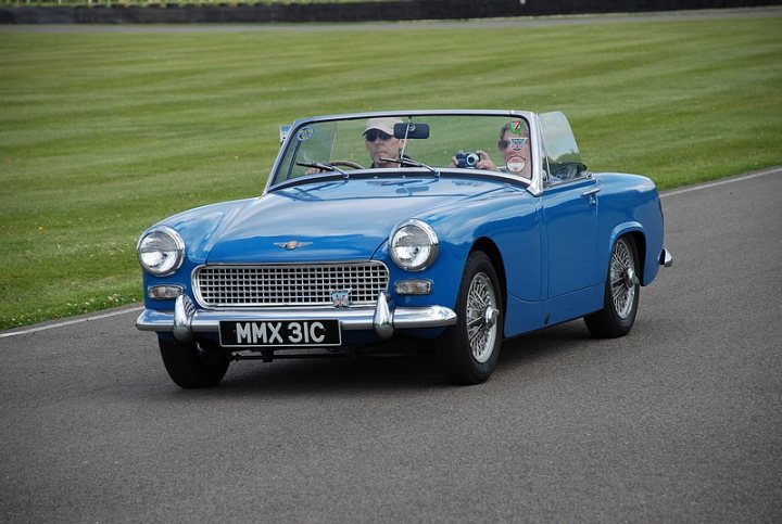 Goodwood Breakfast Club, Pre 1966 classic sunday - Page 1 - Goodwood Events - PistonHeads - The image is a high-resolution photograph featuring a two-door classic blue convertible parked on solid gray pavement. The car has emblems and text on the front, including the model "MMX," denoting year 2000, and the name "EMX," which might be part of a series name. There are two occupants in the vehicle; the one visible is wearing a cap and sunglasses, suggesting a focus on comfort and visibility. The car is situated on the edge of a grassy area, and there is a clear line of sight, focusing the viewer on the convertible and its immediate vicinity.