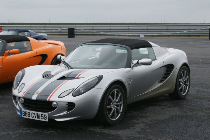 French Photos - Page 1 - Elise/Exige/Europa/340R - PistonHeads - In the image, we see a scene from a race track. The main focus is a striking grey and white Porsche sports car, its sleek body gleaming under the outdoor lighting. Adjacent to it, a vibrant orange sports car is parked on the same track. Both cars, despite their contrasting colors, exude a sense of speed and power. In the backdrop, metal fencing stretches across the image, adding to the racetrack ambiance. The entire scene paints a dynamic picture of a day at the races.