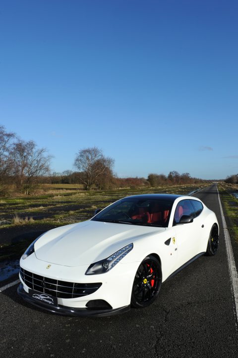 Tragic Ferrari FF.... - Page 1 - Ferrari V12 - PistonHeads - The image features a modern white luxury car captured from an angle that centers the vehicle on a paved road. The car has a sleek design with a bold red stripe running along the side, a distinctive headlight design, and high-performance tires on black alloy wheels. The backdrop of the scene includes a calm, clear day with a few leafless trees indicating a change of seasons, suggesting the photo might have been taken in autumn or winter. The road itself seems to be in the countryside or on the edge of a rural area, as there are no other vehicles or pedestrians visible. The lighting in the image is bright and even, indicating a clear, sunny day.