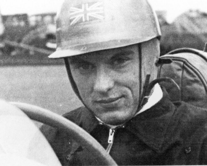 Did Bernie ever drive in Formula 1 - Page 1 - Formula 1 - PistonHeads - The image is a black-and-white photograph featuring a man driving a car. He is wearing a helmet adorned with a Union Jack emblem, indicating a British or British-influenced setting or identity. His attire includes a jacket and possibly a scarf, suggesting a setting that could be colder or dusty, typical of a racing environment. The steering wheel and the patch of a barrier or fence can be seen in the background, verifying the context of a driver inside a moving vehicle.