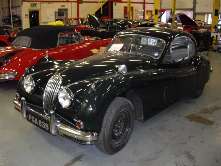 Tour Pistonheads Workshop - The image displays a vintage car in a showroom setting. The car is a classic convertible model, painted in black with silver wire wheels. It features two large round headlights and a large front grille. The vehicle is parked next to a red car and is accompanied by other classic cars in the background. The interior of the showroom includes shelving and storage spaces, suggesting that this is a professional car dealer or exhibition space. The atmosphere suggests a place where automotive enthusiasts can appreciate and possibly acquire such vintage models.