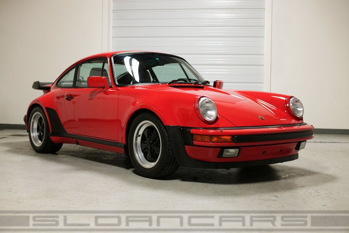 Which car best sums up the 1980s? - Page 7 - General Gassing - PistonHeads - The image displays a vintage red Porsche parked in a garage. The car is predominantly red over a black front strut bar and black wheels with silver hubcaps. The vehicle has a distinctive angular front bumper design, and the image features a watermark. The setting suggests a private collection or display area, with a shutter in the background and no other vehicles visible in the vicinity.