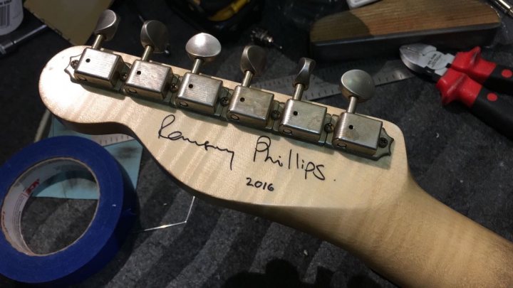 Lets look at our guitars thread. - Page 270 - Music - PistonHeads - The image showcases a close-up of a wooden electric guitar, focusing on its neck area fully visible in the photo. A signature is elegantly written across the length of the neck, indicating ownership or endorsement by someone named Raffaele Phillips. A clear tape, possibly for sealing or marking, partially wraps around the headstock of the guitar. Beside the guitar, a pair of scissors rests, hinting at a possible ongoing project or recent adjustments to the guitar. This scene is set against a dark surface, which might be a workbench, providing a stark contrast that accentuates the details of the guitar and the tools.