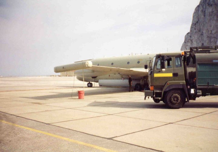 Pistonheads - The image captures a scene on a runway. An old military airplane with propellers is prominently featured in the center. Behind it stands a large six-wheeled truck, suggesting maintenance or transport operations. The tarmac is vast and seems to be deserted apart from a single individual who appears small in comparison to the airplane and truck. In the background, a large rock formation adds a striking natural element to the scene.