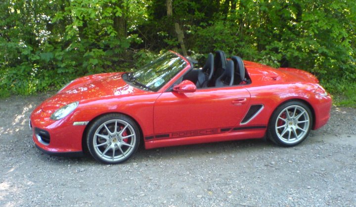 Spyder - help and info please for a prospective new Owner - Page 2 - Porsche General - PistonHeads - The image shows a red Porsche sports car, a classic and often sought-after model, parked amidst a natural setting on a sunny day. The car is close-up, emphasizing its vibrant color and design, with the rear drag from the 911 model visible on the fender. The surrounding area is wooded, with the lush greenery suggesting a peaceful rural area or perhaps a semi-remote subset of the suburban landscape. The lighting and shadows on the white-speckled ground hint at the time of day being either morning or late afternoon.