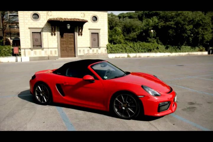 GT4 colours  - Page 52 - Boxster/Cayman - PistonHeads - The image features a vibrant red Porsche sports car parked in an empty parking lot. The car is positioned at an angle to the scene, with its side profile visible, showcasing the sleek design and aerodynamic curves of the Porsche. Surrounding the car is a clear view of a lush green landscape, with numerous trees and shrubs providing a natural setting for the vehicle. On the far left side of the image, a portion of a stone building with arched windows can be seen, which appears to be a larger structure in the background relative to the parked car. The overall scene suggests a peaceful setting, possibly on the outskirts of a town or in a park-like area.