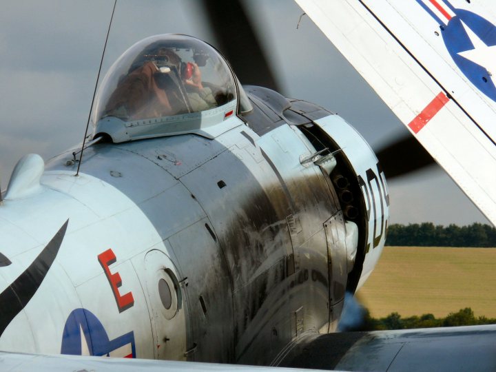 Duxford Images Flying Pistonheads Legends - The image captures a poignant scene: an airplane in flight, passing by in the background. The plane is an older model, possibly a fighter aircraft, given its pointed nose. A distinctive feature is the star emblem on its tail, hinting at a navy affiliation or a flying squadron. Inside, a pilot is visible through the cockpit window, focusing intently on the task at hand. The plane's weathered appearance adds a touch of nostalgia to the image.