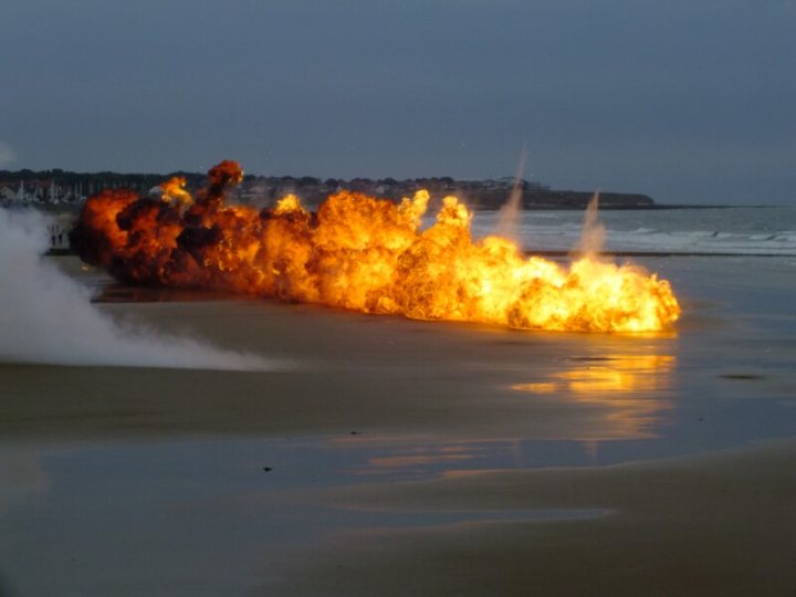 Pistonheads - The image depicts a dramatic scene of a large explosion taking place on a beach. The fireball is bright orange and is surrounded by a darker, cloudier area, possibly the aftermath of the explosion. Smoke and debris are visible around the explosion site, indicating the powerful impact of the blast. The beach stretches out in the background, with dark structures resembling buildings or rocks lining the horizon. The natural lighting suggests that this event may be occurring during the evening or nighttime hours.