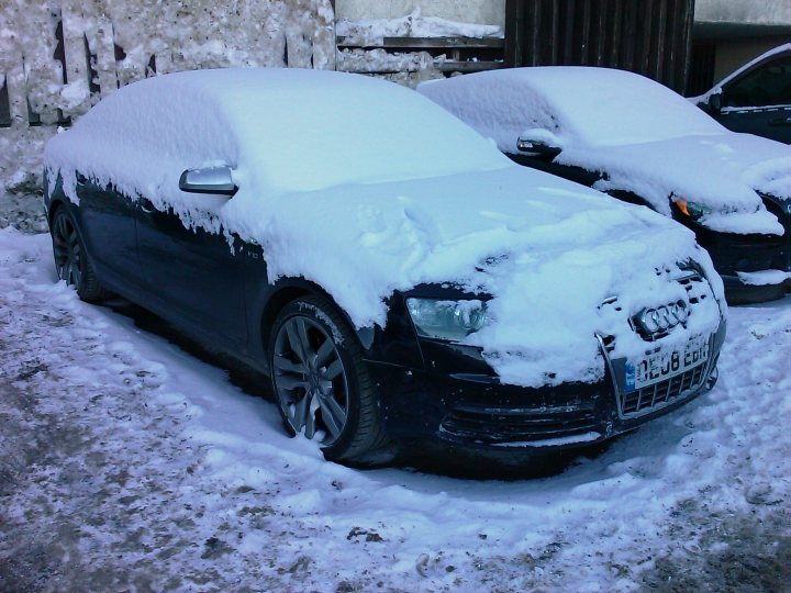 Supercars Dirty Pistonheads - The image shows a sleek, black car parked on a city street. The car is covered in a thick layer of snow, indicating recent heavy snowfall. Another similar car can be seen in the background on the right side of the image. The street appears to be residential, with houses and possibly a fence visible. The overall scene suggests a city experiencing winter weather conditions.