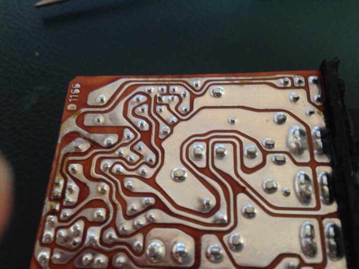 Electronic circuit board / relay repairs - Page 1 - Home Mechanics - PistonHeads - The image is a close-up of a circuit board with numerous electronic components such as resistors and possibly capacitors or other integrated circuits. The board has orange traces connecting various components, indicating electrical connectivity. The circuit board appears small, possibly integrated into a larger system or device. The surface of the board is metallic, and a person's hand can be seen in the top left corner, providing a sense of scale. The image is taken at an angle, allowing the viewer to see two sides of the circuit board simultaneously.