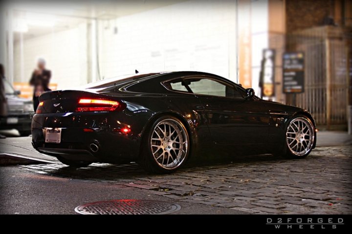 Car Porn - Page 1 - Aston Martin - PistonHeads - The image features an elegant black sports car parked on a cobblestone street. The car is sleek and modern, boasting a glossy finish that gleams under the dusky light. It has large, multi-spoke alloy wheels and a low profile, sitting low to the ground, indicative of a high-performance vehicle. The surroundings include a building, a parking meter, and a partial view of a person standing at a distance, which adds depth to the urban setting. The overall atmosphere of the photo is moody and atmospheric, with a warm, diffused light that suggests either early morning or late evening hours.