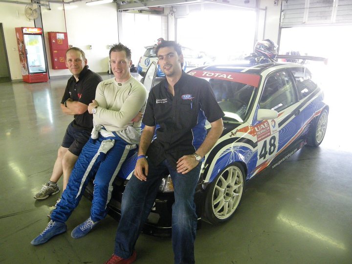 Pistonheads - The image captures a scene in a large garage or workshop. Three men are seated together on the left side, all dressed in casual attire with one noticeably wearing a watch. To the right, a part of a striking blue and white racing car is visible, adorned with a label that reads "48." The car is positioned on a green floor, and the overall atmosphere suggests a gathering or event related to the sport of racing.