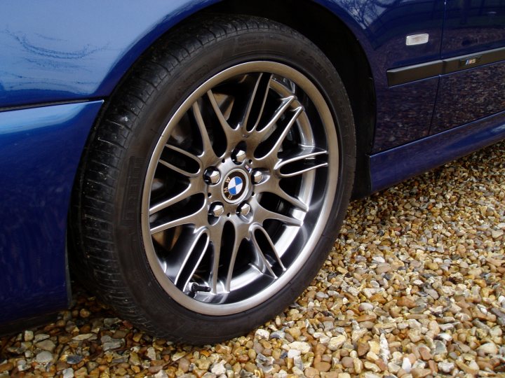 Pistonheads - The image shows a close-up view of a black tire mounted on a silver or white hubcap, with the vehicle positioned on a gravel surface. The tire appears to have a modern design with five spokes that radiate from the center. The hubcap has the BMW logo and additional text, likely including the brand's name. The gravel underneath gives a stark contrast to the smooth and precise finish of the alloy wheel, emphasizing the cleanliness and compactness of the base metal.