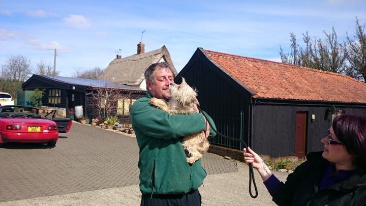 Pistonheads - The image shows a man holding a small cream-colored dog in front of him. The man is dressed in a green jacket and is holding the dog tightly, suggesting a strong bond between them. They seem to be outside, possibly in a driveway, as there are buildings and a car visible in the background. Another person, whose appearance is cropped from the photo, is extending a hand towards the dog. The person appears to be interacting with the dog, possibly petting it. The scene suggests a warm, friendly atmosphere with a focus on the relationship between humans and animals.