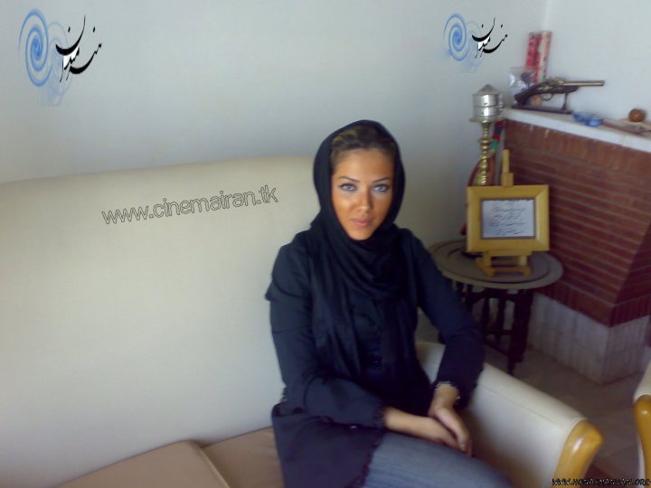 The image shows a woman seated on a beige couch. She is wearing a hijab and is dressed in a black top and black pants. Her eyes are directed toward the camera. In the background, there's a display of Arabic calligraphy, a decorative lamp, and a small sculpture on a pink pedestal. The wall behind has Arabic graffiti on it.