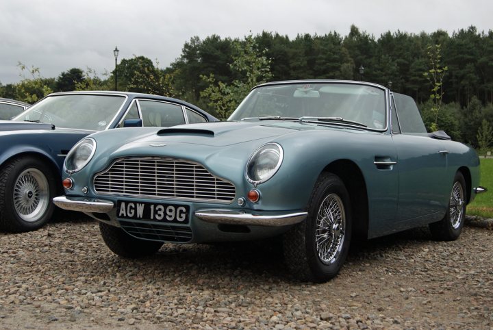 Astons & Ferraris at Glenfiddich distillery - Page 1 - Aston Martin - PistonHeads - The image showcases a vintage Aston Martin car parked outdoors on a gravel surface. The car's body is painted in a metallic green color, matching the backdrop of the surrounding wooded area. The vehicle has a sleek design with a distinctive round front, typical of classic sports cars. The year 1939 is prominently displayed on the rear of the car, suggesting it's a pre-war model. A notable feature is the presence of a white license plate, adding to the authenticity of the image.