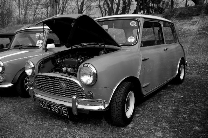 Brooklands Pistonheads Day Mini - The image is in black and white, showcasing two classic Mini Cooper cars. One of the cars has its hood propped open, revealing its engine, which is labeled "KC 587E." The cars are parked in a gravel lot, and there are trees in the background. The image captures the timeless charm of these small, classic vehicles.