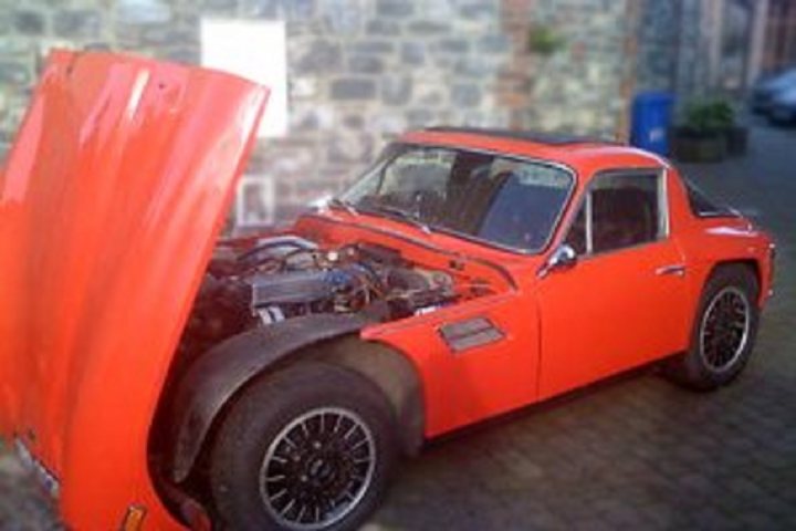 Early TVR Pictures - Page 110 - Classics - PistonHeads - The image shows an orange sports car with the hood propped up, revealing its engine. The car is parked outdoors, possibly on a street, with a brick building and a green plant in the background. There are no people visible in the scene. The car's design suggests it may be a vintage or classic sports car, while the brick wall indicates an urban or semi-urban setting. The green plant adds a touch of nature to the urban environment.