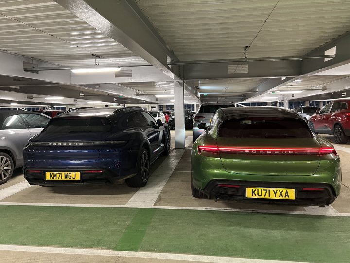 The image shows a parking lot with two rows of cars parked side by side. In the foreground, there are two cars that appear to be luxury models, one green and one blue, positioned on the left side of the photo. On the right side, there is a row of cars, including another luxury vehicle, which looks like a sports car due to its sleek design and the visible spoiler at the rear. The background features a structure with what appears to be parking spaces above, suggesting this could be an indoor or covered parking area.