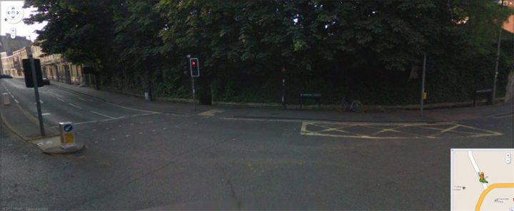 Pistonheads - This is a street view in a calm residential area. The road is curving and unoccupied, with a series of traffic lights visible along the curb. Surrounding the street are lush green trees, adding a natural feel to the scene. To the side of the road, benches can be seen, suggesting a spot for pedestrians to rest. The overhead map image indicates the view is from a vehicle equipped with a navigation system.