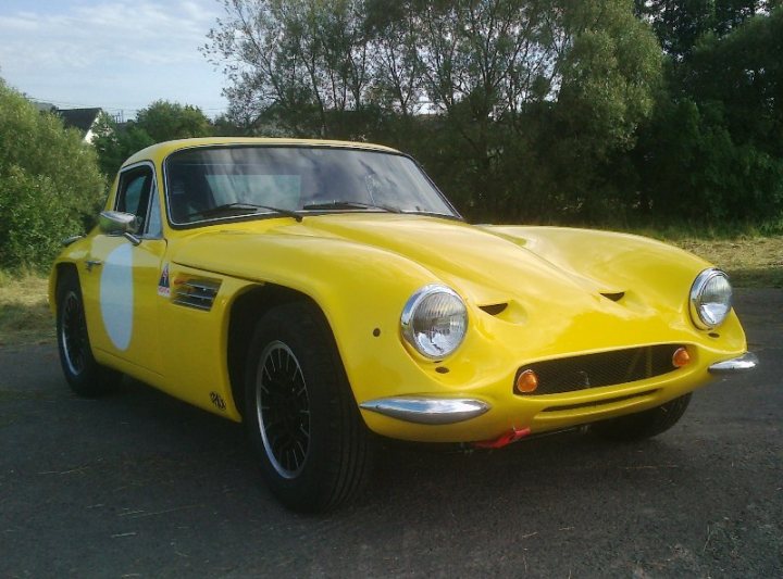 Early TVR Pictures - Page 35 - Classics - PistonHeads - The image showcases a vibrant, yellow vintage car parked on a blacktop road lined with greenery. The car, which appears to have some spots on its body, has a large nose, reminiscent of classic automobile designs, and a grill that features sleek, round headlights. The vehicle's body is adorned with several stripes and a stripe running along the door. The road and surroundings suggest a serene, suburban setting.