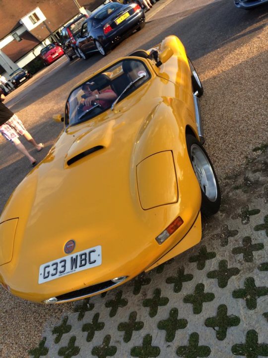 TVRCC Berkshire July meet - Page 1 - Thames Valley & Surrey - PistonHeads - Depicted in the image is a striking yellow sports car parked on a driveway amidst a casual setting. A man, standing in the background, seems oblivious to the car's eye-catching presence. The car and its owner appear to be the primary subjects of the image, indicating that they are the main focus. The backdrop of the scene further accentuates the vibrant car.