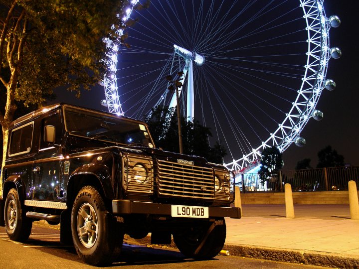 Pistonheads Place Town Night - The image shows a black SUV parked on the side of a sidewalk at night beneath a Ferris wheel. The wheel glows brightly against the dark sky, creating a stark contrast with the vehicle's shiny body. The SUV displays a UK license plate reading L90 MVB, providing a detail about the specific vehicle or its registered owner. There are also cones visible on the side of the vehicle, suggesting it might be associated with an event or some form of construction activity in the area.