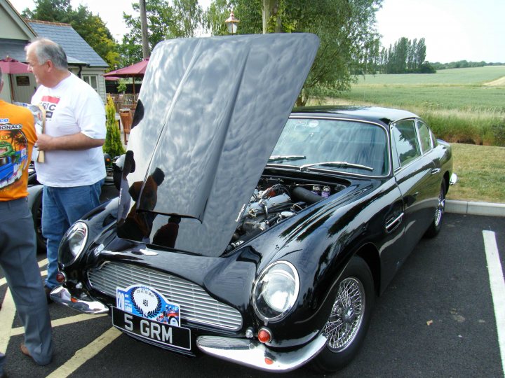 Breakfast Meeting, The Hare, Roxwell 4th June - Page 1 - Kent & Essex - PistonHeads - The image captures a scene in a parking lot where a vintage black car stands out. The car is vintage and black, taking up a large part of the frame. Its hood is open, revealing the intricate engine parts visible under the hood liner. The car is parked on the side of the lot, under the shade of a tree. In the background, two people can be spotted, engaged in conversation, contributing to the overall casual ambiance of the scene.