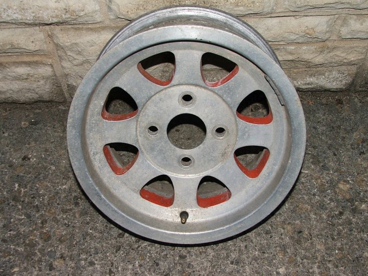 Pistonheads Classic Wheels Identify - The image shows a round metal wheel with a red inner detail, placed next to a textured gray wall, likely made of stone or concrete block. The wheel features a distinct design with holes cut out at various points, suggesting it could be a hubcap or part of a vehicle's steering. The edge of the wheel is flared, and there are visible bolts, possibly indicating its attached components. The wheel is sitting directly on the ground, not supported by any other objects. There is daylight illuminating the scene, casting shadows and highlighting the wheel's texture and design.