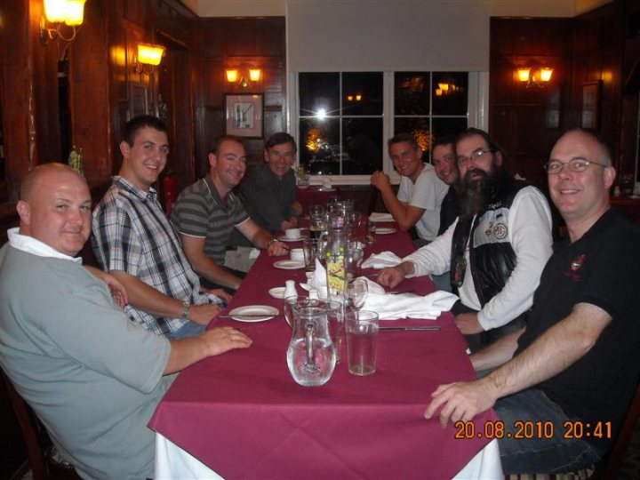 Pistonheads - The image shows a group of eight men seated around a dining table in what appears to be a restaurant. They are all casually dressed and seem to be enjoying a meal together. The table is set with various tableware items including cups, wine glasses, and cutlery. There is also a vase towards the center of the table, which adds a decorative touch to the setting. The men are looking towards the camera, suggesting they may be posing for the photograph.