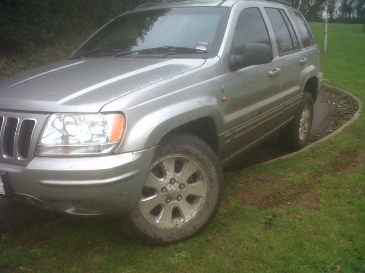 Grand Cherokee, any good? - Page 1 - Off Road - PistonHeads - The image depicts a gray SUV parked on a grassy area with a patch of dirt visible on the ground, suggesting the vehicle has been parked there for some time. The car's design is typical of SUVs, with a raised suspension and robust features. The tires appear to be in poor condition, with visible mud, indicating off-road use or driving on a wet or muddy surface. There is no visible text or distinctive branding on the vehicle that would identify the make or model. The setting seems to be a rural or lightly wooded environment, based on the grass and absence of urban infrastructure.