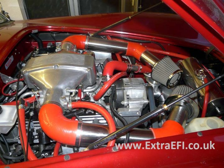 Sequential Fuel and Ignition on 5.0L - Page 1 - Major Mods - PistonHeads - The image shows an inside view of the engine bay of a red car. Various engine components are visible, including a cyclone air intake, red exhaust pipes, and possibly a carburetor and spark plug. The engine is topped by a red wire leading to the fuel pump. The car is indoors, illuminated by artificial lighting, and appears to be in the process of being worked on or maintained, possibly at a repair shop like Extra EF. The photo is a high-quality, close-up shot focused on the intricate parts of the car's engine.