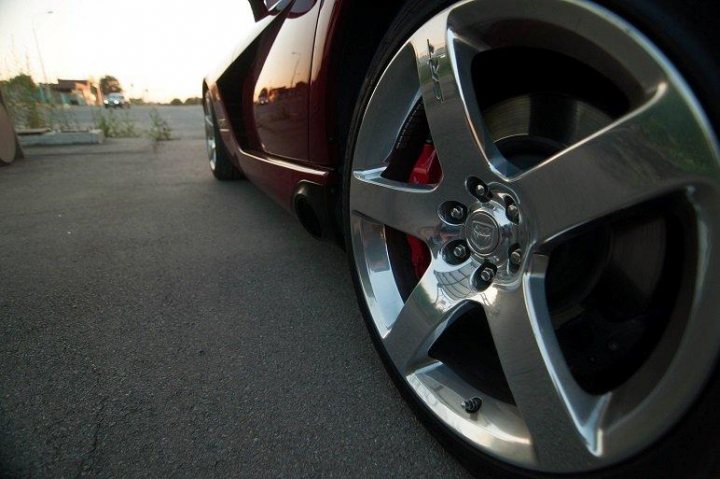 A car is parked next to a parking meter - Pistonheads - The image presents a close-up view of a shiny, black sports car with bright red brakes. The car is parked on a street, showcasing a five-spoke wheel with a silver hub and black rims. The image captures the front right wheel, emphasizing its design and the vehicle's sleek exterior. The setting, mostly out of focus, gives a sense of a quiet city or town area, further highlighting the car.