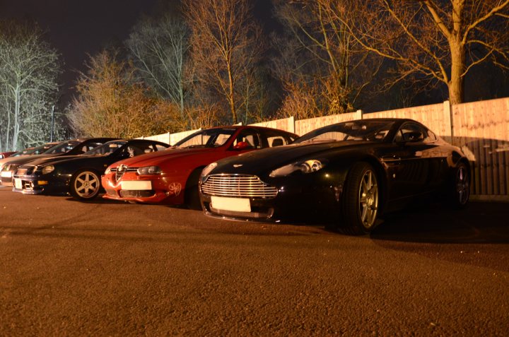 Essex March Pistonheads - The image captures a tranquil nighttime parking area. In the foreground, three luxury sports cars are parked side by side, exhibiting a vibrant blend of red and black. The black topassts of the cars and their sleek, curved shapes add to their glossy appearance. The red car's convertible top is open, revealing a black interior.

The cars are parked on a dark asphalt parking lot, adjacent to a wooden fence. The wooden fence separates the parking area from a lush landscape of trees under the starlit sky. The contrast between the dark vehicles and the illuminated parking lot creates a captivating scene, highlighting the elegance of the cars and the serene surroundings.