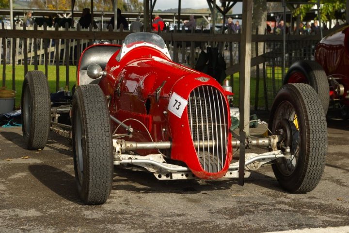 Pistonheads - The image captures a row of vintage cars, each distinct in its design, parked side by side in an outdoor setting. They're situated in front of a wooden fence, adding a rustic charm to the scene. The sun casts shadows on the ground and the cars, indicating an open-air parking area, possibly of a classic vehicle show. The cars' gleaming surfaces reflect the sunlight, enhancing their vintage appeal. The nearest car is a striking red sports car from the number 26, its presence dominating the foreground of the image.