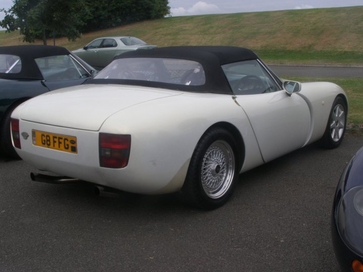 Griff Pistonheads Growl - The image shows a vintage sports car parked in an outdoor lot. The car has a sleek white exterior with black trim, giving it an elegant and sporty appearance. It features a black convertible top and a distinctive yellow license plate with black lettering. The vehicle is positioned at an angle facing towards the left of the frame, providing a clear view of its front and side profile. The lot is bordered by a grassy area and appears to be a public parking space, as another car is parked to the left of the vintage sports car.