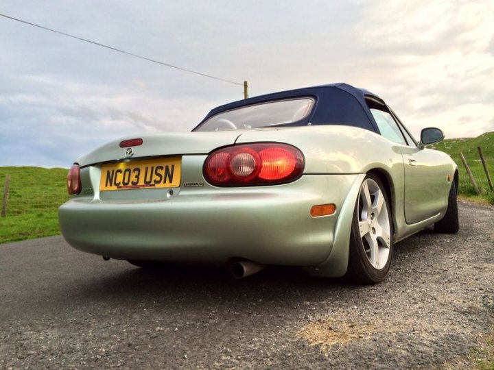 my mx5 - Page 1 - Mazda MX5/Eunos/Miata - PistonHeads - The image depicts a silver two-door convertible sports car parked on a gravel road with grassy hills visible in the background. The car displays a U.K. license plate with the letters "NCO3 USN" suggesting a UK-based military connection. The rear feature of the car is a prominent set of vertical taillights, which is characteristic of some European vehicle designs. The vehicle appears to be in good condition, and the sun's reflections suggest it might be a sunny day. The noise reduction in the image is notably absent, as evidenced by the clear and uncut text on the license plate.