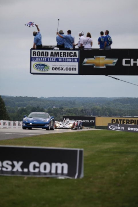 Pistonheads - The image captures an exciting moment in a road race. There are several people on top of a bridge, standing next to the sign that reads "Road America Elkhart Lake, WI OSX.com". They all appear to be enthusiastic spectators, possibly posing for a photo or simply enjoying the race. 

Below the bridge, there is a vibrant scene of the race in progress. Two race cars are in the foreground, one leading the other, both exhibiting the speed and agility typical for such events. 

The bridge has a metal guard rail, suggesting it's designed to withstand the high volumes of vehicles passing underneath during the race. 

The bridge and spectators are positioned against a backdrop of lush green trees and a clear blue sky, providing a picturesque setting for this exhilarating event.